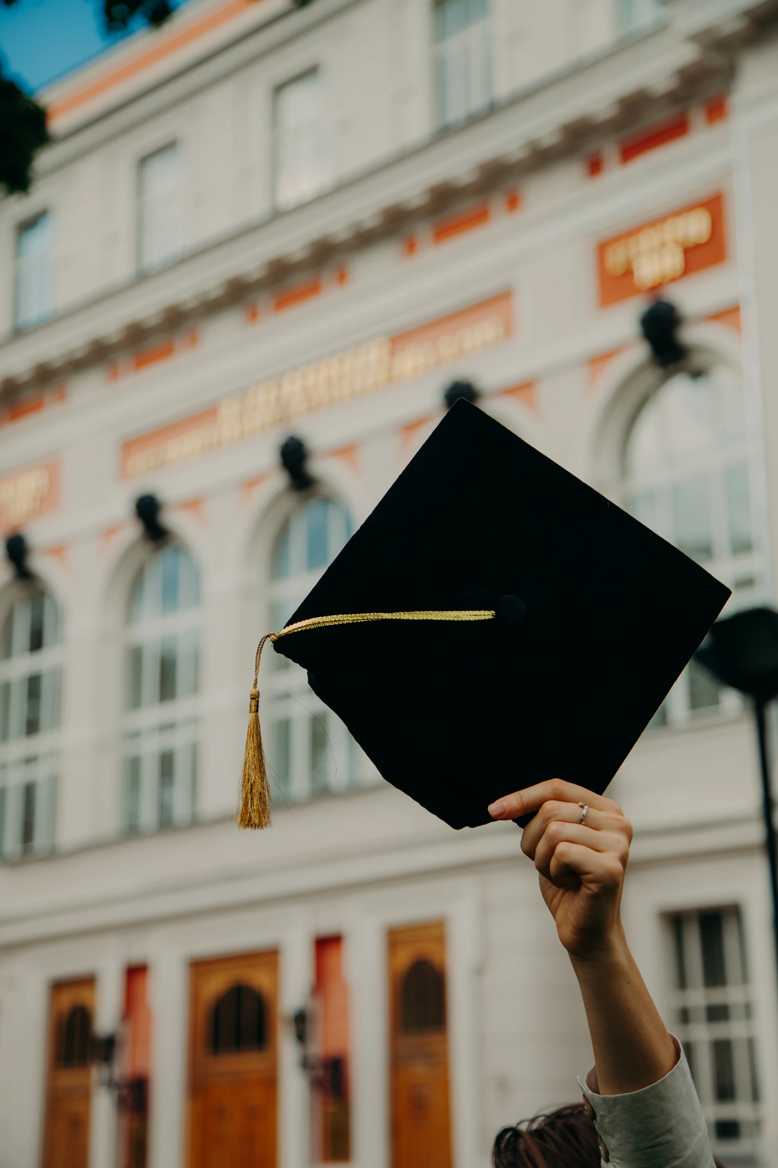 A hand holding up a black graduation cap in front of a historic building