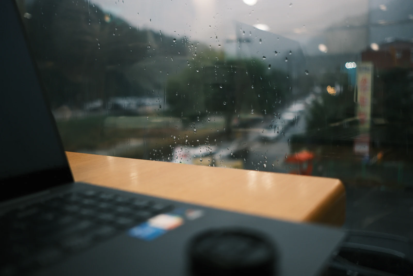 A laptop on a wooden desk beside a window streaked with rain