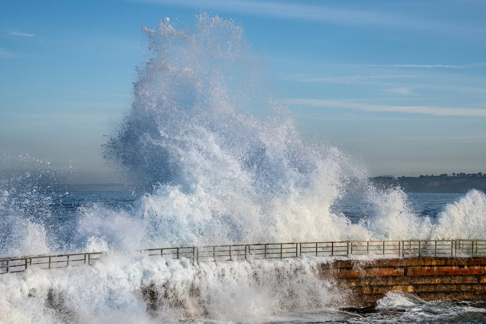 A large wave crashing over a stone sea wall with metal railings