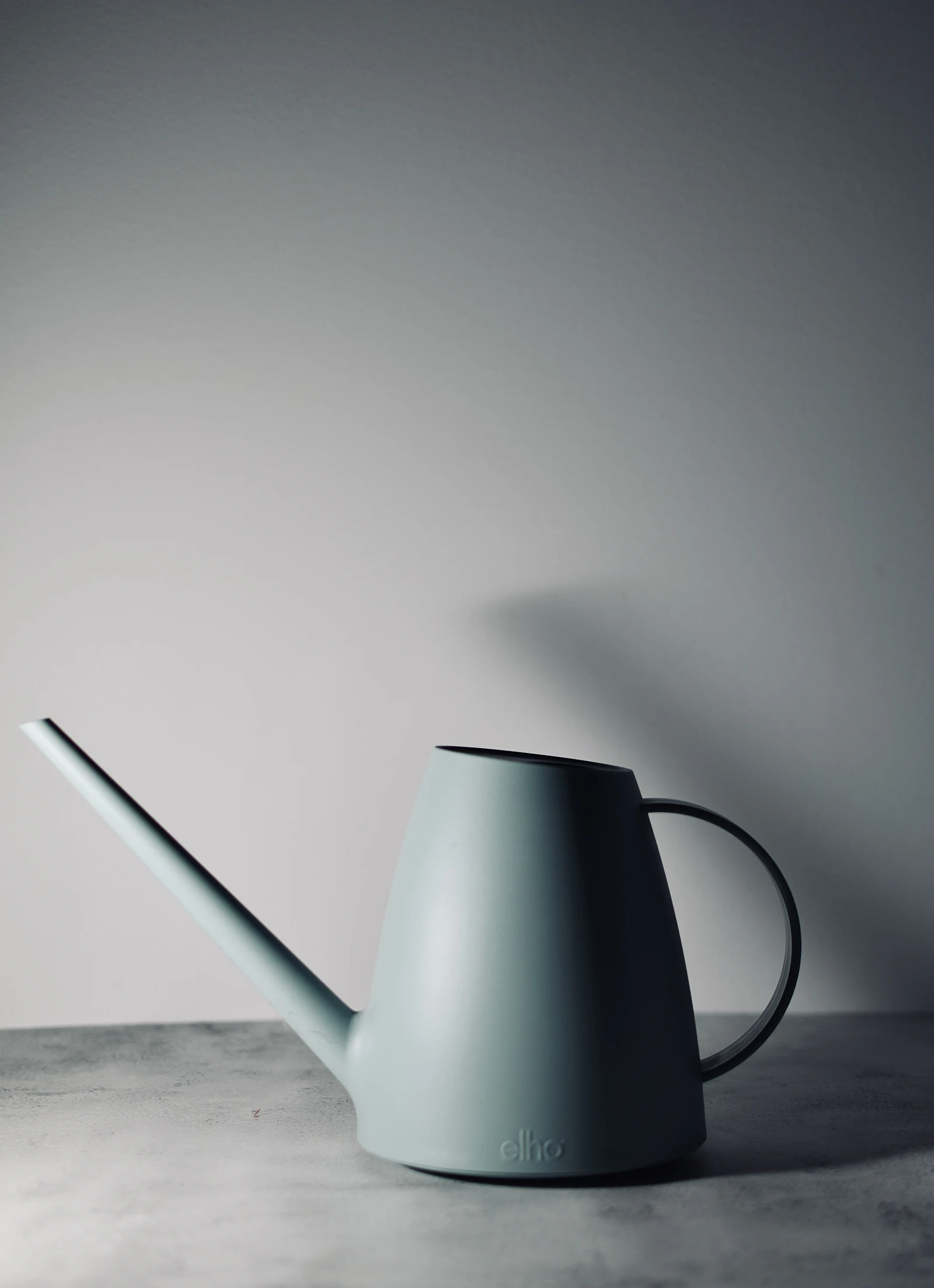 A grey watering can with a curved handle and long spout against a grey wall